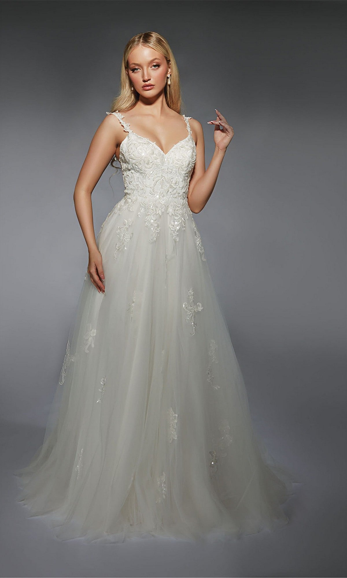 A woman wearing the Long White Dress: Alyce 7124, a floor-length A-line gown with delicate lace appliques, stands against a plain gray background.